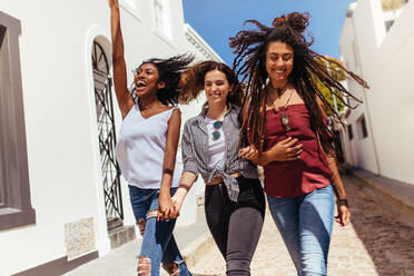 Drei glücklich aussehende Frauen, die an einem sonnigen Tag auf der Straße spazieren gehen und modische Kleidung tragen. Junge Frau springt vor Freude, während sie mit Freunden auf einer Straße spazieren geht. - JLPSF26676