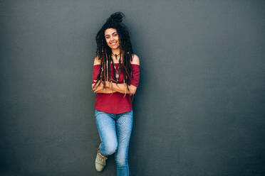 Porträt einer jungen Frau mit lockigem Haar, die modische Kleidung und Accessoires trägt und an einer Wand steht. Lächelnde Frau, die mit verschränkten Armen an einer Wand steht. - JLPSF26678