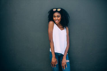 Portrait of a curly haired woman wearing fashionable clothes standing against a wall. Smiling woman standing against a wall wearing sunglasses on her head. - JLPSF26680