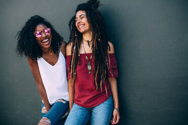 Zwei Frauen in modischer Kleidung stehen an einer Wand. Glücklich aussehende Frauen stehen an einer Wand und tragen modische Accessoires und Sonnenbrillen. - JLPSF26686