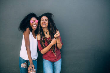 Zwei Frauen in modischer Kleidung stehen an einer Wand. Glücklich aussehende Frauen stehen an einer Wand und tragen modische Accessoires und Sonnenbrillen. - JLPSF26687