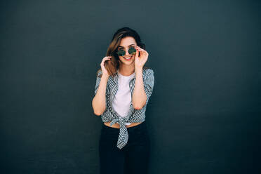 Lächelnde Frau mit modischer Kleidung und Sonnenbrille, die an einer Wand steht. Glücklich aussehende Frau, die an einer Wand steht und für ein Foto posiert. - JLPSF26690