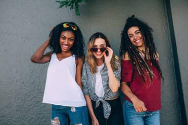 Drei Frauen in modischer Kleidung stehen im Freien an einer Wand. Glücklich aussehende Frauen stehen an einer Wand und posieren für ein Foto. - JLPSF26694