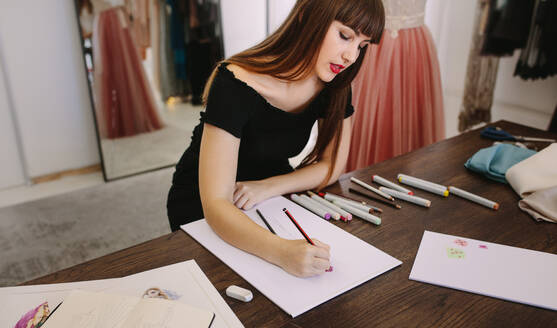 Modeunternehmerin, die an ihrem Tisch sitzend einen Entwurf skizziert. Modedesignerin, die in ihrem Stoffgeschäft eine Zeichnung anfertigt. - JLPSF26696