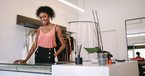 Weibliche Modedesignerin steht an ihrem Schreibtisch in ihrer Boutique. Weibliche Kleiderdesignerin in ihrem Stoffladen mit ausgestellter Designerkleidung. - JLPSF26709