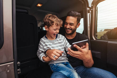 Aufnahme eines glücklichen Vaters und eines Sohnes, die auf der Rückbank des Autos sitzen und sich mit einem digitalen Tablet amüsieren. Ein junger Mann und ein kleiner Junge lächeln mit einem digitalen Tablet. - JLPSF26718
