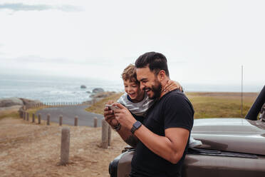 Outdoor shot of happy father and son in front of the car using mobile phone and laughing. Young man and little boy watching something on smart phone and enjoying while on road trip. - JLPSF26720