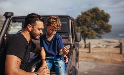 Kleiner Junge mit Smartphone auf der Motorhaube sitzend mit seinem Vater. Vater und Sohn vor dem Auto mit Handy. - JLPSF26721