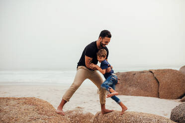 Shot of father and son on the beach vacation. Young man playing with little boy at the sea shore. Jumping on the rocks. - JLPSF26727