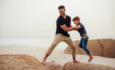 Aufnahme eines Vaters, der seinem Sohn hilft, über einen Felsen am Strand zu springen. Ein junger Mann und ein kleiner Junge haben Spaß am felsigen Strand. - JLPSF26728