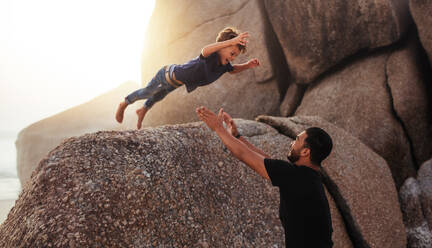 Aufnahme eines süßen kleinen Jungen, der von einem großen Felsen in die Arme seines Vaters springt. Vater und Sohn genießen die Sommerferien am Strand. - JLPSF26730