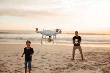 Vater und Sohn bedienen die Drohne per Fernsteuerung am Meer. Junger Mann mit kleinem Jungen fliegt Drohne am Strand während des Sommerurlaubs. Fokus auf Drohne, mit Vater und Sohn im Hintergrund. - JLPSF26742