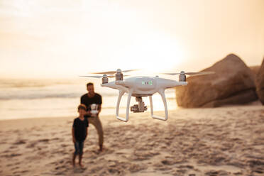 Drohne wird von Vater und Sohn am Meer betrieben. Vater und Sohn im Sommerurlaub fliegen Drohne am Strand. Fokus auf Drohne, mit Vater und Sohn im Hintergrund. - JLPSF26743