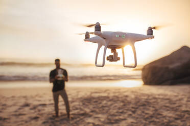 Drohne wird von einem Mann bedient, der im Hintergrund am Strand steht. Junger Mann am Meeresufer, der eine Drohne fliegt. - JLPSF26744