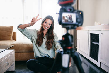 Young woman vlogger showing victory or peace sign while recording her daily video blog. Vlogger using a camera mounted on a tripod to record her video. - JLPSF26751