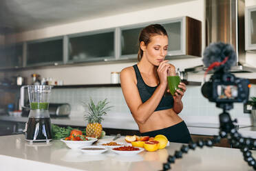 Junge Frau in der Küche trinkt gesunden Gemüsesaft, während sie ihn mit der Kamera aufnimmt. Obst und Gemüse zum Frühstück auf dem Küchentisch. - JLPSF26757