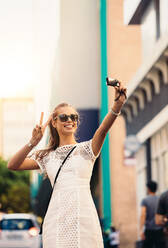 Young woman posing for a selfie in a street flashing victory sign. Vlogger recording content for her travel vlog. - JLPSF26767