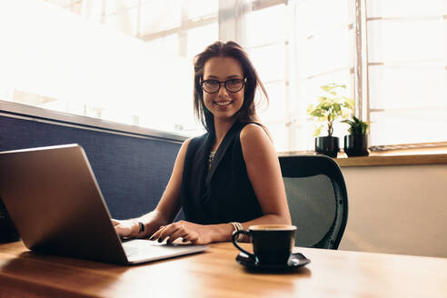 Weibliche Vloggerin, die ihren Vlog am Computer bearbeitet. Lächelnde junge Frau an ihrem Schreibtisch im Büro, die mit einer Tasse Kaffee am Computer arbeitet. - JLPSF26768