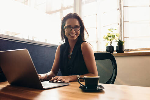 Weibliche Vloggerin, die ihren Vlog am Computer bearbeitet. Lächelnde junge Frau an ihrem Schreibtisch im Büro, die mit einer Tasse Kaffee am Computer arbeitet. - JLPSF26769