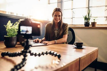 Junge Frau, die ein Video für ihren Vlog mit einer Digitalkamera auf einem flexiblen Stativ aufnimmt. Lächelnde Frau, die an ihrem Schreibtisch sitzt und an einem Laptop arbeitet. - JLPSF26773