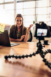 Junge Frau, die ein Video für ihren Vlog mit einer Digitalkamera auf einem flexiblen Stativ aufnimmt. Lächelnde Frau, die an ihrem Schreibtisch sitzt und an einem Laptop arbeitet und in die Kamera schaut. - JLPSF26774
