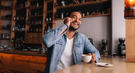 Innenaufnahme eines lächelnden jungen Mannes, der in einem Café sitzt und mit einem Mobiltelefon spricht. - JLPSF26805