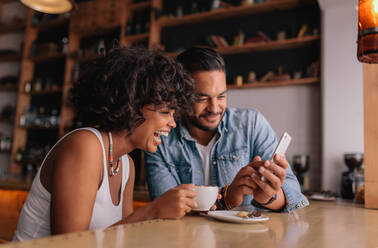 Glückliches junges Paar, das in einem Café sitzt, auf sein Handy schaut und lächelt. Junger Mann und Frau in einem Café, die ein Smartphone benutzen. - JLPSF26809