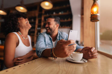 Junges Pärchen in einem Café, ein Mann hält ein Handy in der Hand und eine lachende Frau sitzt daneben. - JLPSF26814