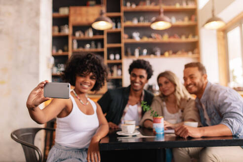 Eine gemischtrassige Gruppe von Freunden macht ein Selfie mit ihrem Smartphone in einem Café. Junge Männer und Frauen sitzen an einem Cafétisch und machen ein Selbstporträt mit ihrem Mobiltelefon - JLPSF26818