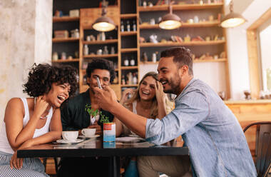 Junge Gruppe von Freunden, die in einem Café sitzen und ein Mobiltelefon benutzen. Junger Mann und Frauen sitzen am Cafétisch und lachen. - JLPSF26819
