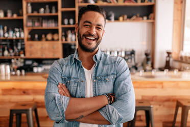 Porträt eines glücklichen jungen Mannes, der mit verschränkten Armen in einem Café steht. Lächelnder gut aussehender Mann, der in einem Café steht. - JLPSF26823