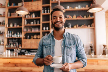 Porträt eines gutaussehenden jungen Mannes, der mit einer Tasse Kaffee in einem Café steht. Lächelnder junger Mann beim Kaffeetrinken in einem Restaurant. - JLPSF26824