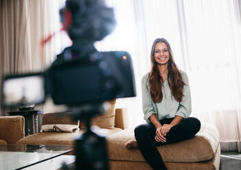 Junge Frau, die ein Video mit einer auf einem Stativ montierten Kamera für ihren Vlog aufnimmt. Hübsche Frau, die in die Kamera lächelt, während sie in ihrem Wohnzimmer sitzt. - JLPSF26828