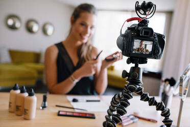 Eine Frau macht ein Video für ihren Blog über Kosmetika. Eine junge Frau mit Kosmetikartikeln auf einem Tisch nimmt ihr Video mit einer auf einem Stativ montierten Kamera auf. - JLPSF26833