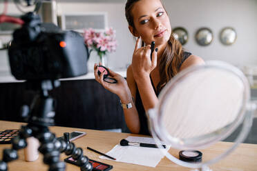 Modebloggerin, die ein Video für ihren Blog über Kosmetik aufnimmt. Junge Frau, die Make-up aufträgt und in eine auf einem Stativ befestigte Kamera schaut. - JLPSF26834