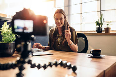 Junge Frau, die ein Video für ihren Vlog mit einer Digitalkamera auf einem flexiblen Stativ aufnimmt. Lächelnde Frau, die an ihrem Schreibtisch sitzt und an einem Laptop arbeitet. - JLPSF26842