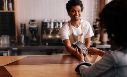 Young woman paying by credit card at cafe. Woman entering security pin in credit card reader with male barista standing behind checkout counter at coffee shop. - JLPSF26852