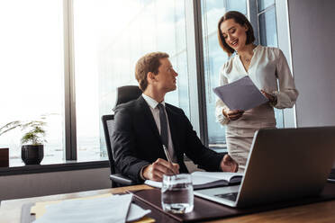 Two businesspeople working together in office. Businessman sitting at desk with his secretary reading notes in a modern office. - JLPSF26858