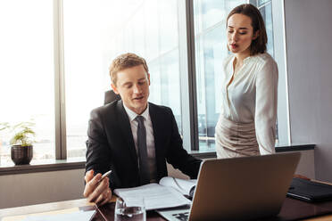 Businessman reading and discussing contract paper terms with female colleague standing by at his desk. Corporate professionals working together in office. - JLPSF26859