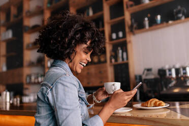 Seitenansicht der lächelnden jungen afrikanischen Frau, die an der Kaffeetheke sitzt, frühstückt und Kaffee trinkt. - JLPSF26860