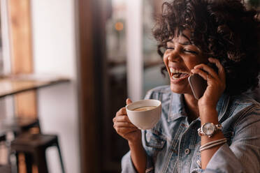 Fröhliche junge Frau telefoniert und lächelt, während sie einen Kaffee in einem Café genießt. Afrikanische Frau benutzt ein Mobiltelefon in einem Café. - JLPSF26861