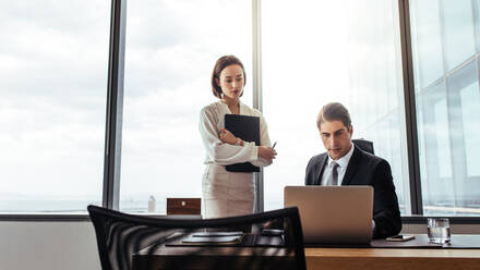 Geschäftsmann, der an einem Laptop arbeitet, mit einer Sekretärin, die daneben steht. Zwei Geschäftsleute arbeiten zusammen im Büro. - JLPSF26862