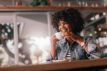 Porträt einer schönen jungen afrikanischen Frau, die in einem Café sitzt und Kaffee trinkt. Lächelnde junge Frau, die in einem Restaurant Kaffee trinkt. - JLPSF26863