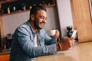 Lächelnder junger kaukasischer Mann, der in einem Café sitzt, sein Handy benutzt und Kaffee trinkt. Gut aussehender Mann mit einer Tasse Kaffee und einer Textnachricht auf seinem Smartphone. - JLPSF26864