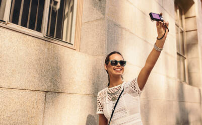 Stylische junge Frau, die mit einer Digitalkamera ein Selfie auf der Straße macht. Mode-Vloggerin, die Inhalte für ihren Vlog aufnimmt. - JLPSF26866