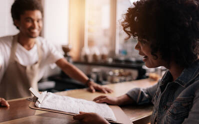 Afrikanische Frau gibt dem Barista, der hinter dem Tresen eines Cafés steht, eine Bestellung auf. Frau bestellt Kaffee am Tresen eines Cafés. - JLPSF26870