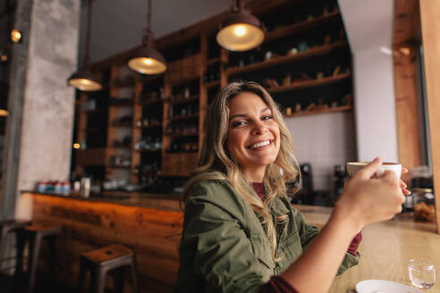 Porträt einer schönen jungen Frau, die mit einer Tasse Kaffee im Café sitzt und lächelt. Kaukasische Frau trinkt Kaffee im Café. - JLPSF26874