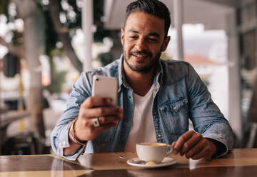 Indoor shot of young man sitting in coffee shop reading text message on mobile phone. Handsome caucasian guy at cafe using smart phone. - JLPSF26878