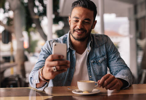 Innenaufnahme eines jungen Mannes, der in einem Café sitzt und eine Textnachricht auf seinem Handy liest. Gut aussehender kaukasischer Mann im Café, der sein Smartphone benutzt. - JLPSF26878