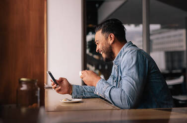 Seitenansicht des glücklichen jungen Mannes, der in einem Café sitzt und sein Handy benutzt. Gutaussehender kaukasischer Mann im Café, der sein Smartphone benutzt und Kaffee trinkt. - JLPSF26881